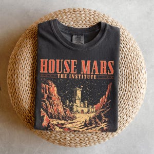 May include: Dark gray t-shirt with the text "HOUSE MARS THE INSTITUTE" in orange. The graphic features a castle scene with red rock formations and a starry night sky. The shirt is folded on a woven surface.