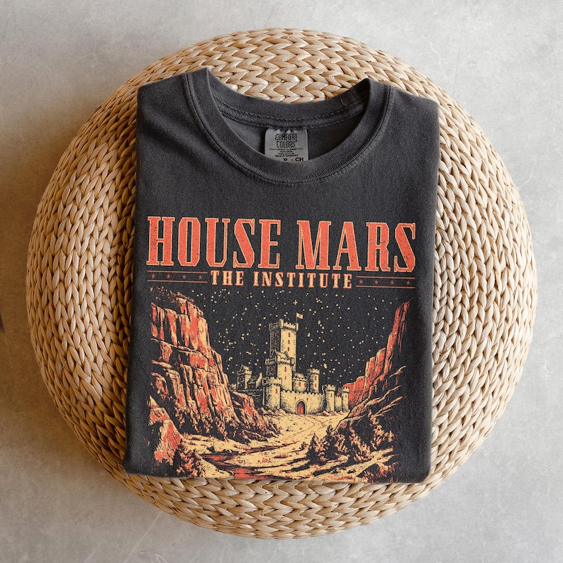 May include: Dark gray t-shirt with the text "HOUSE MARS THE INSTITUTE" in orange. The graphic features a castle scene with red rock formations and a starry night sky. The shirt is folded on a woven surface.