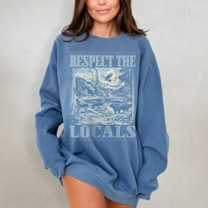 May include: A blue sweatshirt with a vintage-style graphic print of a mountain scene with the text "Respect the Locals".
