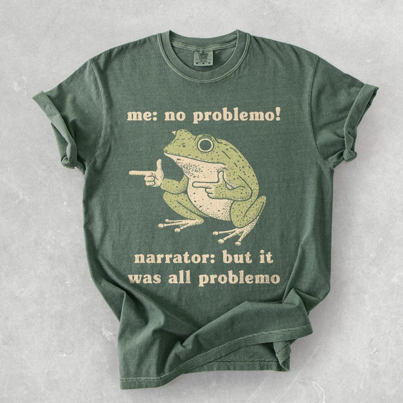May include: Green t-shirt with a frog graphic and the text "me: no problemo!" and "narrator: but it was all problemo". The frog is light green and beige, with a pointing gesture.