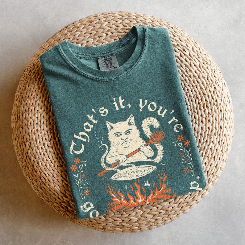 May include: Teal-colored t-shirt with a graphic of a cat cooking over a fire, holding a wooden spoon. The text "That's it, you're going" is arched around the cat illustration. The shirt is folded on a woven, circular mat.