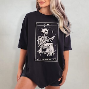 Op de afbeelding: Zwart T-shirt met een tarotkaart ontwerp. De kaart toont een skelet dat een boek leest, omlijst door sterren en manen, met de tekst "THE READER" eronder. Het shirt heeft korte mouwen en een losse pasvorm.