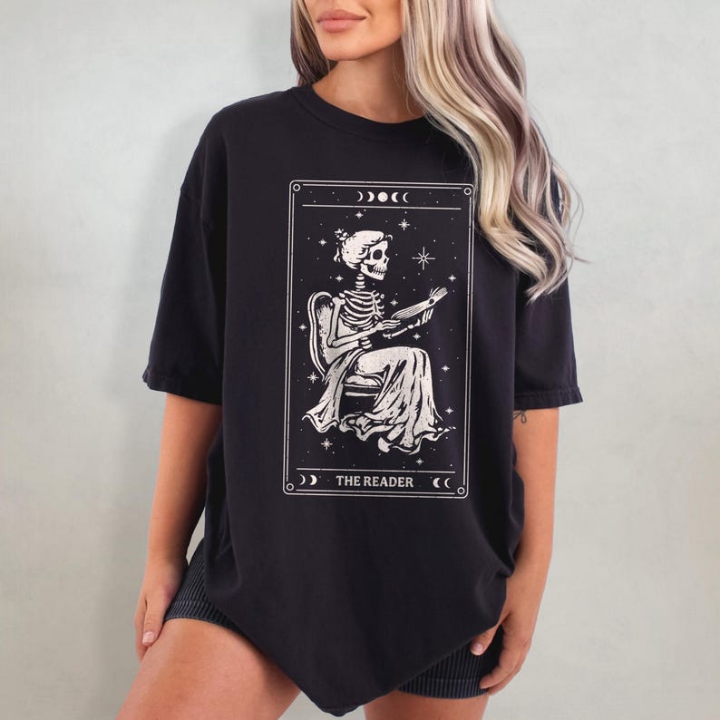 Op de afbeelding: Zwart T-shirt met een tarotkaart ontwerp. De kaart toont een skelet dat een boek leest, omlijst door sterren en manen, met de tekst "THE READER" eronder. Het shirt heeft korte mouwen en een losse pasvorm.