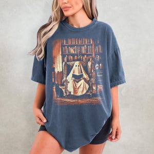 Op de afbeelding: Donkerblauw T-shirt met een afbeelding van een spook dat in een bibliotheek leest. De afbeelding bevat een zwarte kat, boekenplanken en een fauteuil. Het ontwerp heeft een vintage, licht vervaagde uitstraling.