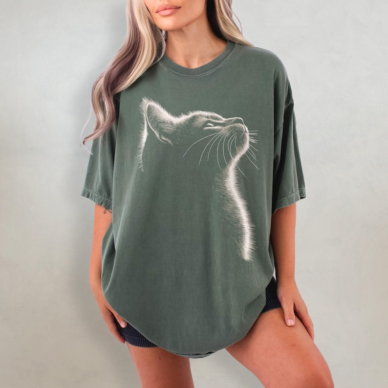 May include: An oversized green t-shirt featuring a white outline of a cat's face looking upwards. The cat's whiskers are visible.