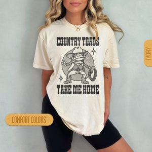 May include: A white t-shirt with a cartoon frog wearing a cowboy hat and holding a lasso. The text "COUNTRY TOADS TAKE ME HOME" is printed on the shirt.