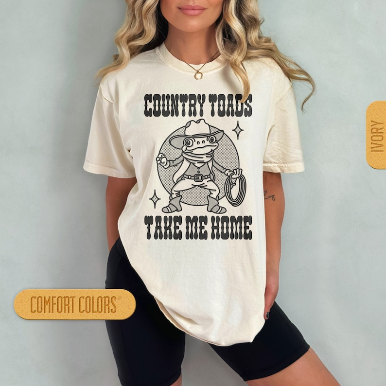 May include: A white t-shirt with a cartoon frog wearing a cowboy hat and holding a lasso. The text "COUNTRY TOADS TAKE ME HOME" is printed on the shirt.