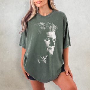May include: An olive green t-shirt featuring a white portrait of a man's face. The short-sleeved shirt has a relaxed fit and a vintage, faded appearance. The graphic is a detailed, grayscale image of a man's profile.