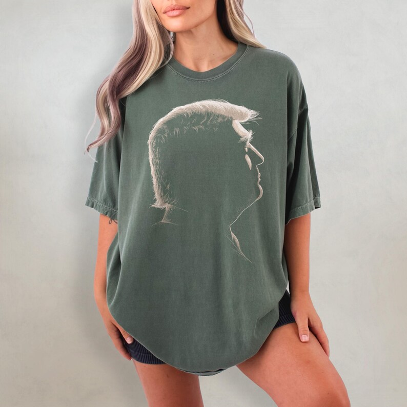 K&ouml;nnte beinhalten: Ein olivgr&uuml;nes T-Shirt mit einer Profilsilhouette einer Person mit wei&szlig;en Haaren. Die Grafik ist auf der Vorderseite zentriert. Das Shirt hat kurze &Auml;rmel und einen Rundhalsausschnitt.