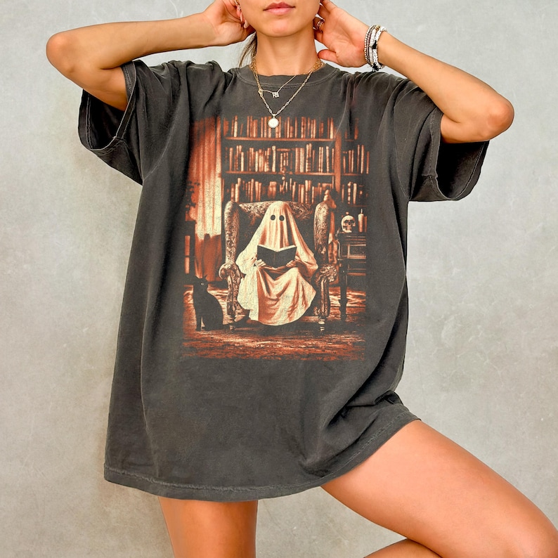 Op de afbeelding: Donkergrijs t-shirt met een afbeelding van een geest die in een bibliotheek leest. Het ontwerp bevat een zwarte kat, boekenplanken en een schedel. Het t-shirt heeft een vintage, versleten uitstraling.