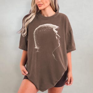K&ouml;nnte beinhalten: Braunes T-Shirt mit einem wei&szlig;en Seitenprofil-Portr&auml;t. Das T-Shirt hat kurze &Auml;rmel und eine lockere Passform. Die Grafik ist eine Silhouette des Kopfes und der Schultern einer Person.