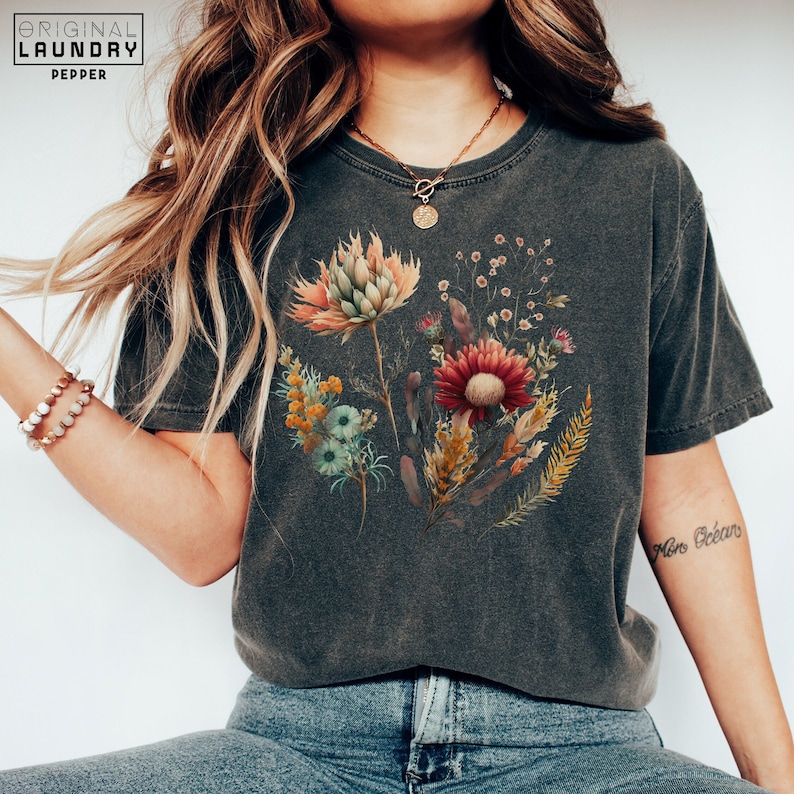 May include: A dark gray t-shirt with a floral print featuring a variety of flowers in shades of red, orange, yellow, and blue. The flowers are arranged in a bouquet-like shape and are set against a distressed background.