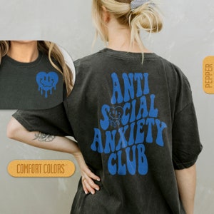 K&ouml;nnte beinhalten: Ein schwarzes T-Shirt mit einem blauen Aufdruck, der "Anti Social Anxiety Club" in einer Retro-Schriftart zeigt. Der Aufdruck enth&auml;lt auch ein blaues, tropfendes Smiley-Gesicht. Das Shirt wird von einem Model getragen.