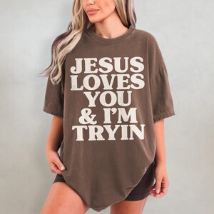May include: A brown oversized t-shirt with the text "JESUS LOVES YOU & I'M TRYIN" printed in white.