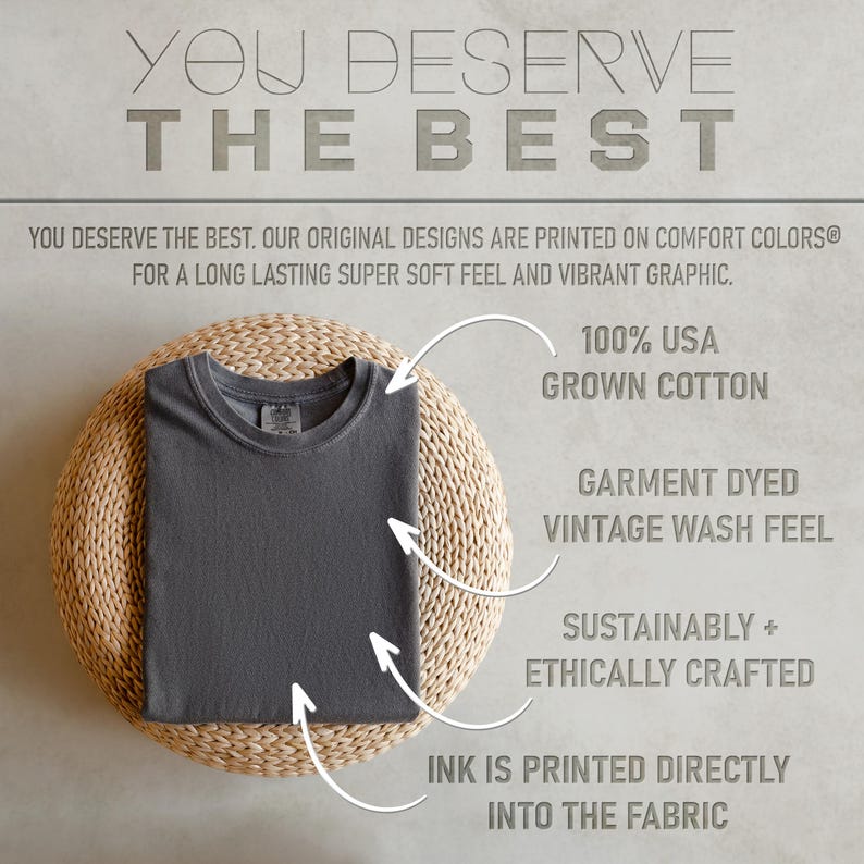 Op de afbeelding: Een gevouwen, antracietgrijs t-shirt ligt op een ronde, geweven ondergrond. De tekst erboven luidt "YOU DESERVE THE BEST." Extra tekst benadrukt kenmerken: 100% in de VS geteeld katoen, geverfd met vintage wassing, duurzaam en ethisch vervaardigd, en inkt direct in de stof gedrukt.