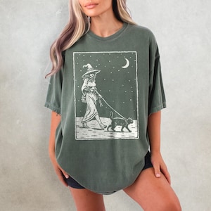 May include: Dark green t-shirt featuring a white graphic of a witch walking a cat under a starry night sky with a crescent moon. The witch is wearing a hat and dress. The cat is on a leash.