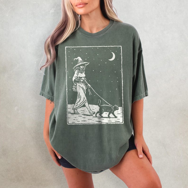 May include: Dark green t-shirt featuring a white graphic of a witch walking a cat under a starry night sky with a crescent moon. The witch is wearing a hat and dress. The cat is on a leash.