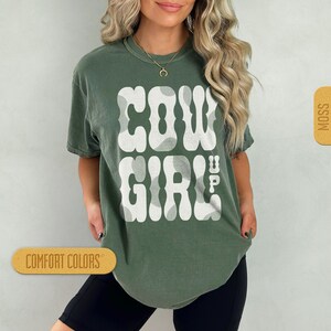 May include: A green t-shirt with a white graphic that says "COWGIRL" with a cow print pattern. The shirt is a relaxed fit and has a crew neck.