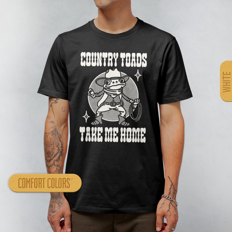 May include: Black t-shirt with a white graphic of a cartoon frog wearing a cowboy hat and holding a lasso. The text "COUNTRY TOADS TAKE ME HOME" is printed in white on the shirt.