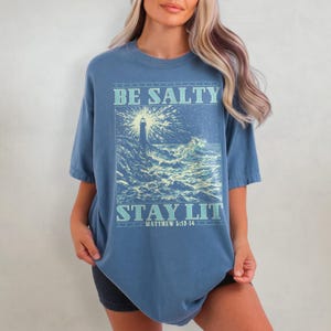 May include: A blue oversized t-shirt with a graphic of a lighthouse in a stormy sea. The text "BE SALTY STAY LIT" is printed on the shirt, with the scripture reference "MATTHEW 5:13-16" underneath.