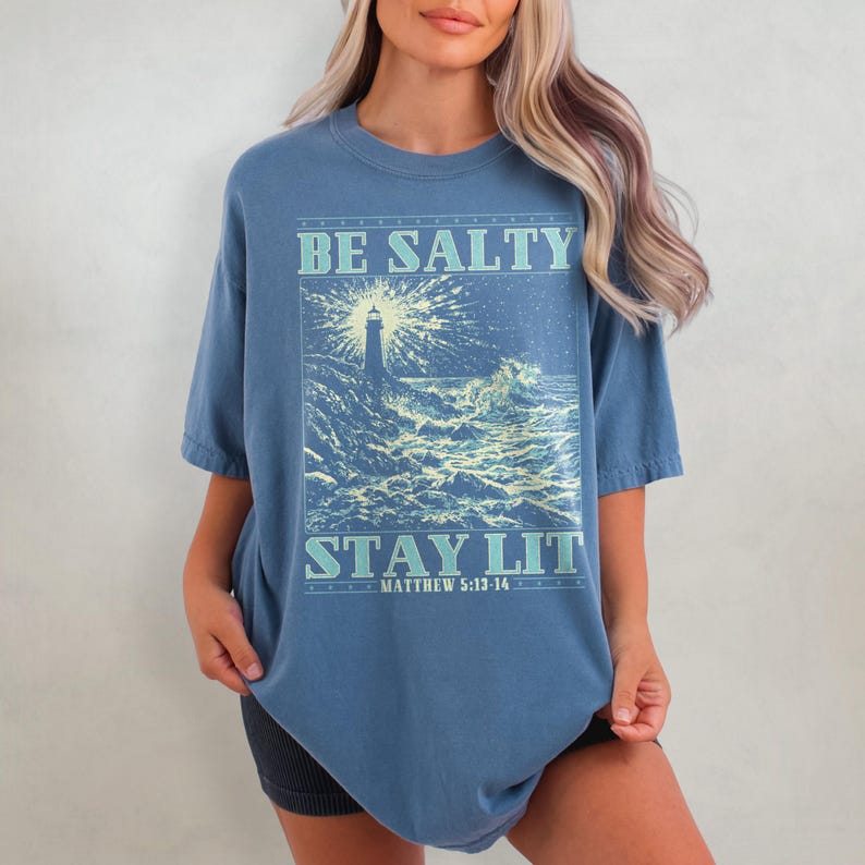 May include: A blue oversized t-shirt with a graphic of a lighthouse in a stormy sea. The text "BE SALTY STAY LIT" is printed on the shirt, with the scripture reference "MATTHEW 5:13-16" underneath.
