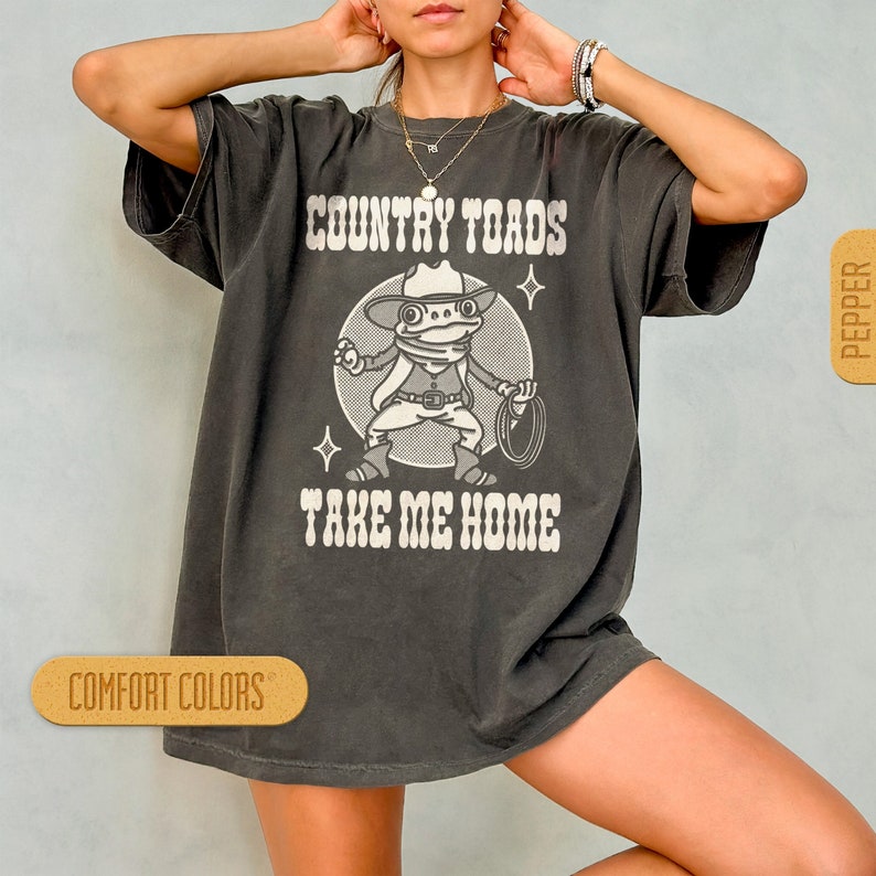 May include: A dark gray t-shirt with a cartoon frog wearing a cowboy hat and holding a lasso. The text "COUNTRY TOADS TAKE ME HOME" is printed on the shirt.