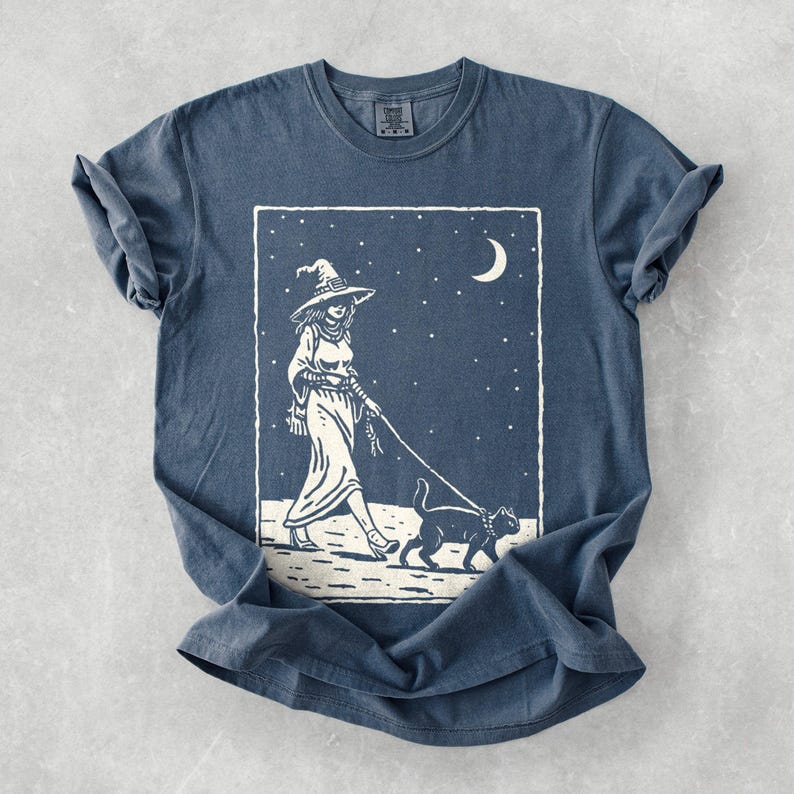 May include: A blue t-shirt featuring a white graphic of a witch walking a black cat under a starry night sky with a crescent moon. The witch is wearing a hat and dress. The shirt is folded.