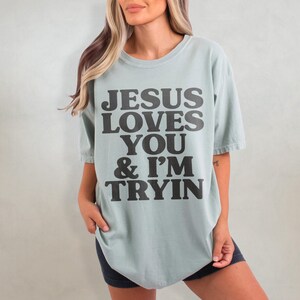 May include: A light blue t-shirt with the text "JESUS LOVES YOU & I'M TRYIN" printed in black.