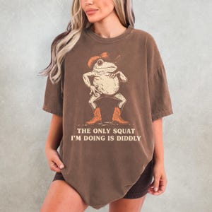 May include: Brown t-shirt featuring a cartoon frog wearing a cowboy hat and boots. The frog is in a squatting pose. The text on the shirt reads, "THE ONLY SQUAT I'M DOING IS DIDDLY."