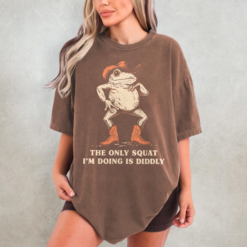 May include: Brown t-shirt featuring a cartoon frog wearing a cowboy hat and boots. The frog is in a squatting pose. The text on the shirt reads, "THE ONLY SQUAT I'M DOING IS DIDDLY."