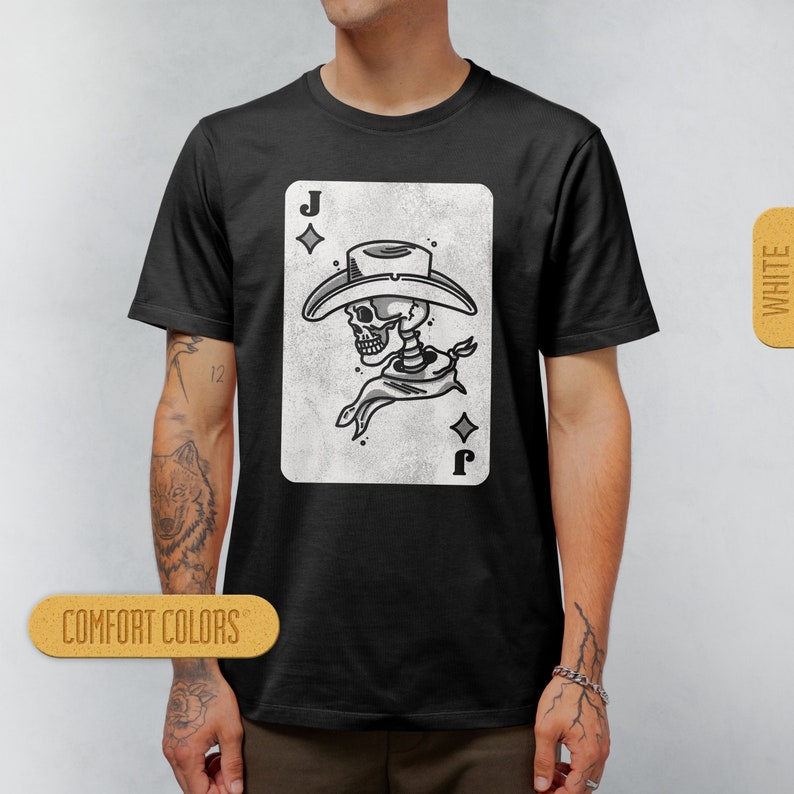 Puede incluir: Camiseta negra con un gr&aacute;fico blanco de un esqueleto con un sombrero de vaquero y un pa&ntilde;uelo. El gr&aacute;fico es una carta de juego con la letra 'J' y un s&iacute;mbolo de diamante. El texto 'COMFORT COLORS' es visible en la parte inferior izquierda de la imagen.