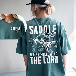 May include: Teal-colored t-shirt with a white graphic of a cowboy riding a horse. The text "SADDLE UP" is above the graphic, and "WE'RE FOLLOWING THE LORD" is below. The shirt has a crew neck and short sleeves.