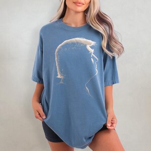 K&ouml;nnte beinhalten: Ein blaues T-Shirt mit einem Seitenprofil einer Person mit hellem Haar. Die Grafik ist eine Silhouette in einem helleren Blauton. Das T-Shirt hat kurze &Auml;rmel und einen Rundhalsausschnitt.