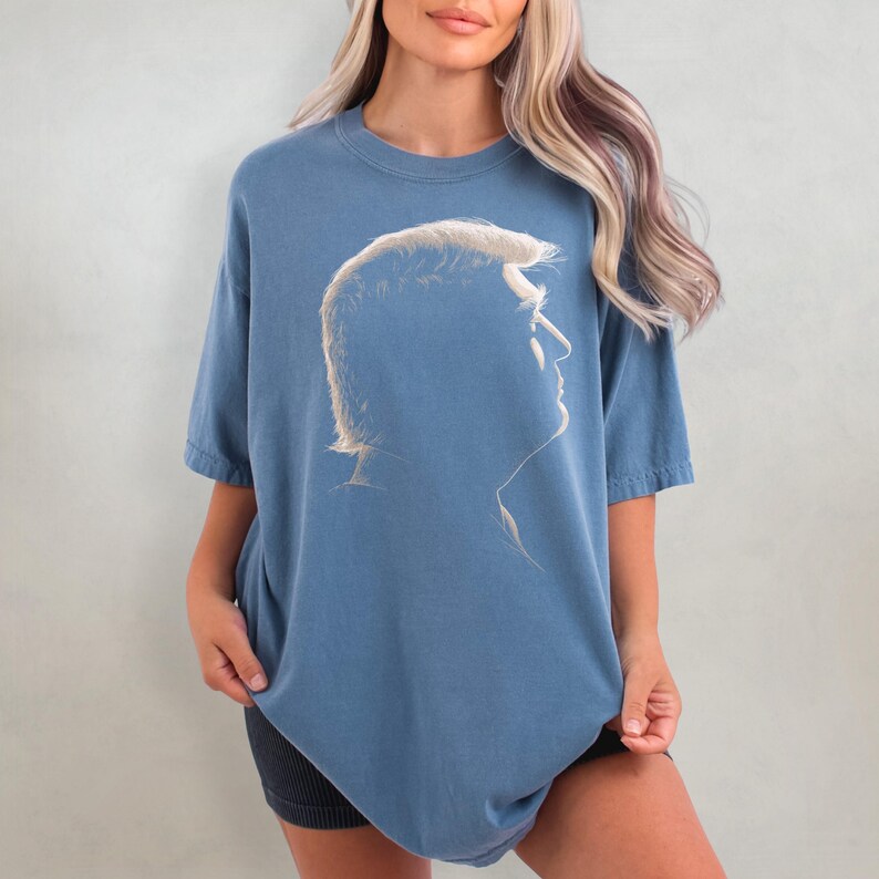 K&ouml;nnte beinhalten: Ein blaues T-Shirt mit einem Seitenprofil einer Person mit hellem Haar. Die Grafik ist eine Silhouette in einem helleren Blauton. Das T-Shirt hat kurze &Auml;rmel und einen Rundhalsausschnitt.