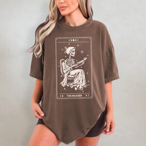 Op de afbeelding: Bruin t-shirt met een tarotkaartontwerp. De afbeelding toont een skelet dat een boek leest, zittend in een stoel, met de tekst "THE READER" onderaan. Het ontwerp is omlijst met sterren en halve manen.