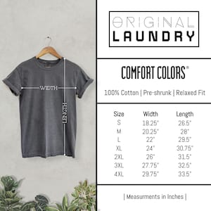 May include: A gray Comfort Colors t-shirt hanging on a wooden hanger. The shirt has a relaxed fit and is made of 100% cotton. Measurements in inches are provided for sizes S through 4XL. The brand name "Original Laundry" is displayed.