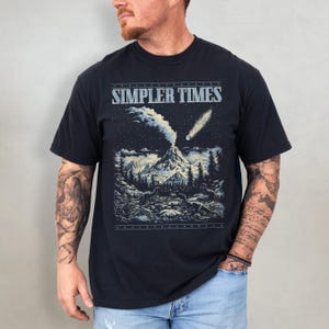 K&ouml;nnte beinhalten: Schwarzes T-Shirt mit dem Text "SIMPLER TIMES" &uuml;ber einer Grafik eines ausbrechenden Vulkans, eines Kometen und Dinosauriern in einem Wald. Das Design ist in Blau- und Wei&szlig;t&ouml;nen gehalten.