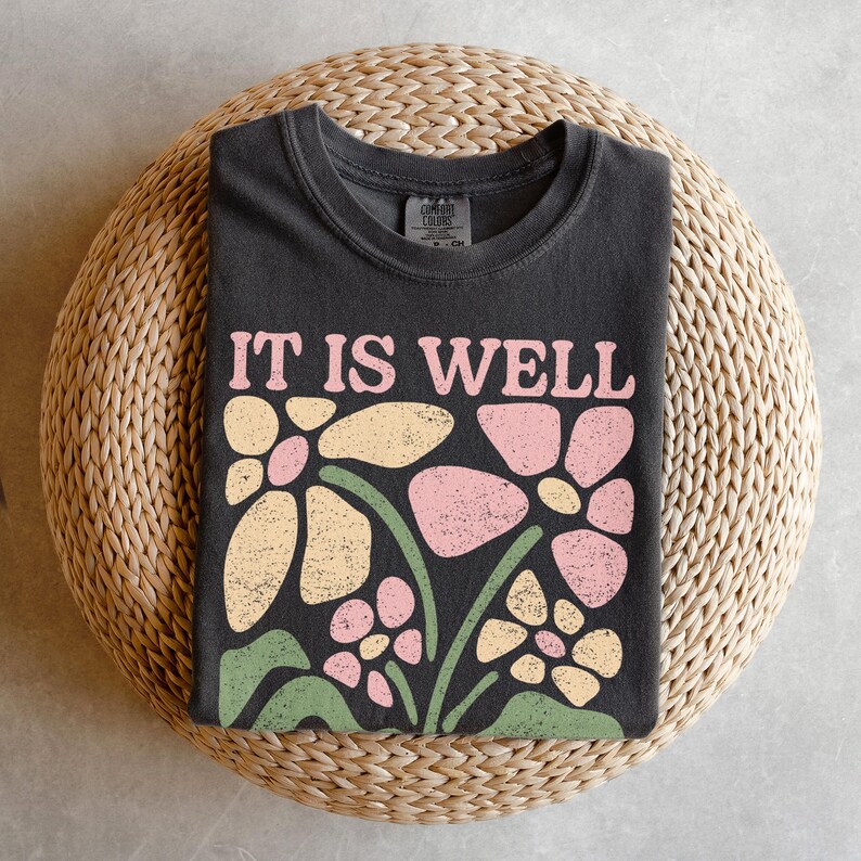 May include: A dark gray t-shirt with the words "IT IS WELL" in pink and cream-colored letters. Below the text is a floral design in pink, cream, and green. The shirt is folded on a woven, circular surface.