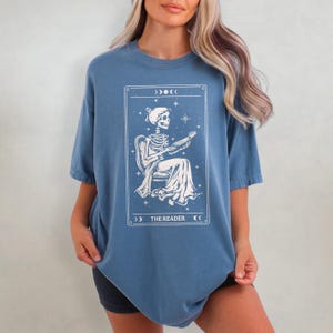 Op de afbeelding: Blauw T-shirt met een witte afbeelding van een skelet dat een boek leest, omlijst door een decoratieve rand met maan- en steraccenten. De tekst "THE READER" staat onderaan de afbeelding.