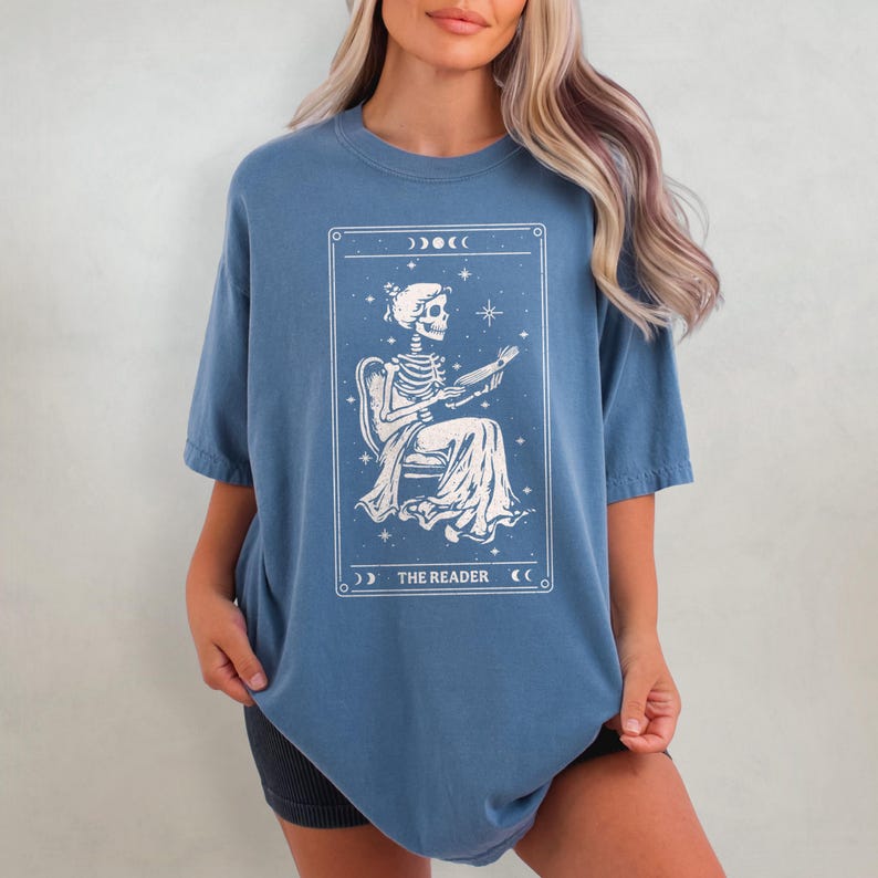 Op de afbeelding: Blauw T-shirt met een witte afbeelding van een skelet dat een boek leest, omlijst door een decoratieve rand met maan- en steraccenten. De tekst "THE READER" staat onderaan de afbeelding.