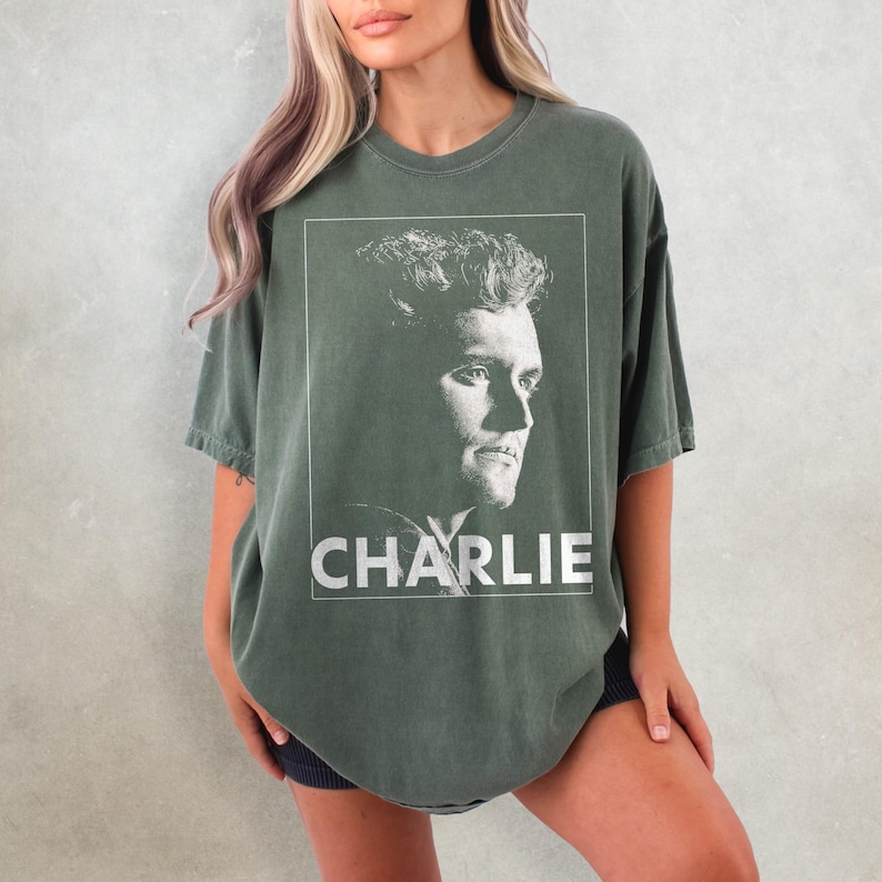 May include: An olive green t-shirt with a black and white portrait of a man in a white frame. The name "CHARLIE" is printed in white block letters below the portrait. The shirt has short sleeves and a relaxed fit.