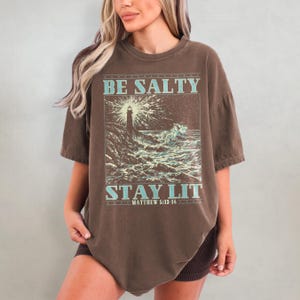 May include: Brown oversized t-shirt with a graphic of a lighthouse and waves. The text "BE SALTY STAY LIT" is printed on the shirt with the scripture reference "MATTHEW 5:13-14" underneath.