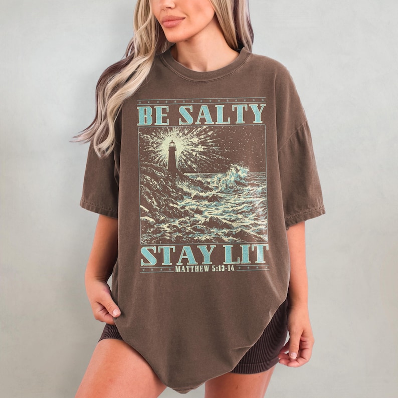 May include: Brown oversized t-shirt with a graphic of a lighthouse and waves. The text "BE SALTY STAY LIT" is printed on the shirt with the scripture reference "MATTHEW 5:13-14" underneath.