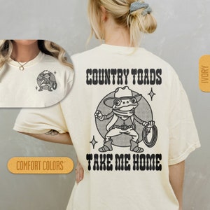 Puede incluir: Una camiseta de color crema con una ilustraci&oacute;n en blanco y negro de una rana con un sombrero de vaquero y sosteniendo un lazo. El texto "COUNTRY TOADS TAKE ME HOME" est&aacute; impreso en negro en la camiseta.