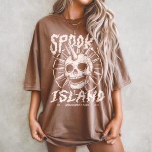 May include: Brown t-shirt with a distressed graphic featuring a skull with a horn, surrounded by spiky rays. The text reads "SPOOK ISLAND AMUSEMENT PARK 2002" in a vintage font. The shirt has short sleeves and a relaxed fit.