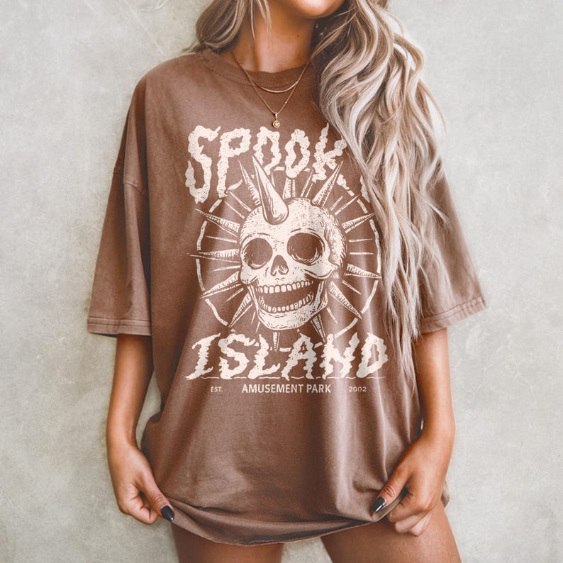 May include: Brown t-shirt with a distressed graphic featuring a skull with a horn, surrounded by spiky rays. The text reads "SPOOK ISLAND AMUSEMENT PARK 2002" in a vintage font. The shirt has short sleeves and a relaxed fit.