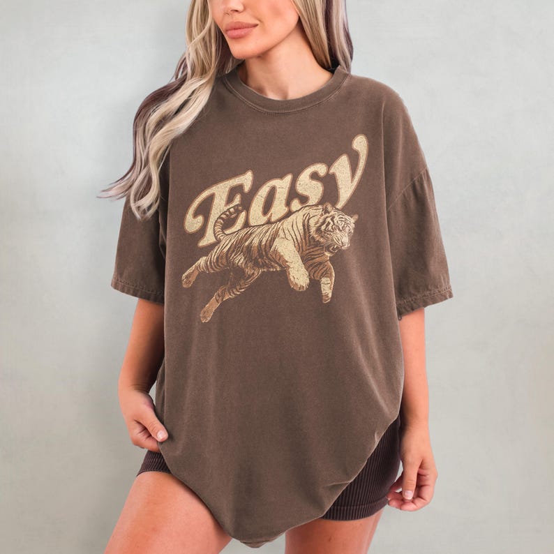Peut inclure: Un t-shirt surdimensionn&eacute; marron avec un motif vintage d'un tigre sautant avec le mot "Easy" dans une police r&eacute;tro.