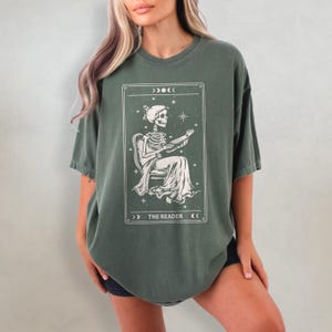 Op de afbeelding: Olijfgroen t-shirt met een tarotkaart ontwerp. De kaart toont een skeletfiguur zittend in een stoel, een boek vasthoudend. De tekst "THE READER" staat onderaan de kaart.