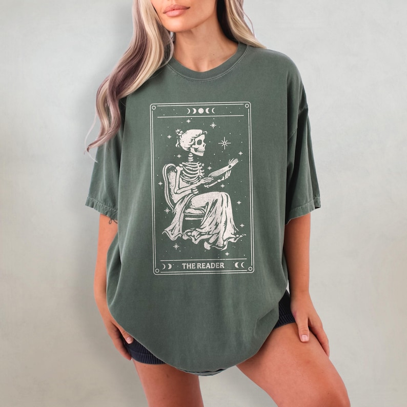 Op de afbeelding: Olijfgroen t-shirt met een tarotkaart ontwerp. De kaart toont een skeletfiguur zittend in een stoel, een boek vasthoudend. De tekst "THE READER" staat onderaan de kaart.