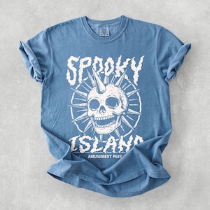 May include: A blue t-shirt with a white graphic featuring a skull with a horn, surrounded by spiky lines. The text "Spooky Island Amusement Park" is printed below the skull in a distressed font.